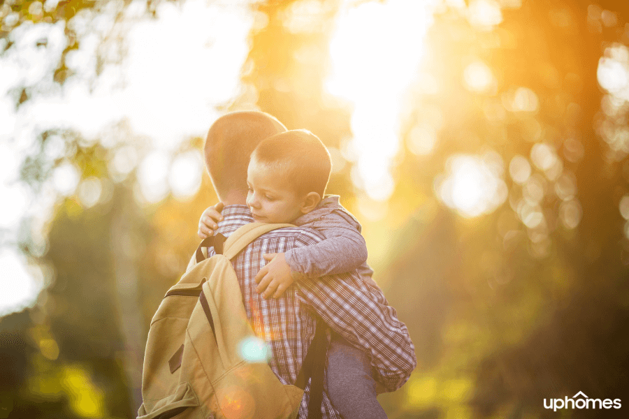 Single Dad with backpack carrying son in his arms with sun shining through to showcase the light at the end of the tunnel