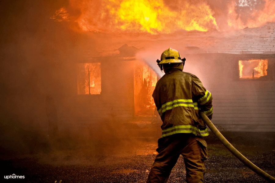 House on fire with a firefighter in the foreground putting out the blaze with a hose and water wearing a firefighter suit