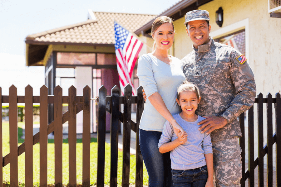 A military service member in uniform standing with their spouse and child in front of a picket fence outside their new home.