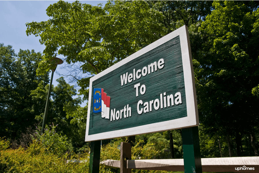A green Welcome to North Carolina sign with the NC State flag and green trees in the background