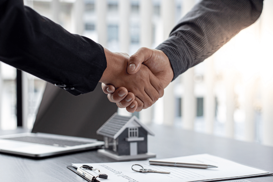 Shaking hands with a laptop, pen, paper, keys, and house replica in the background