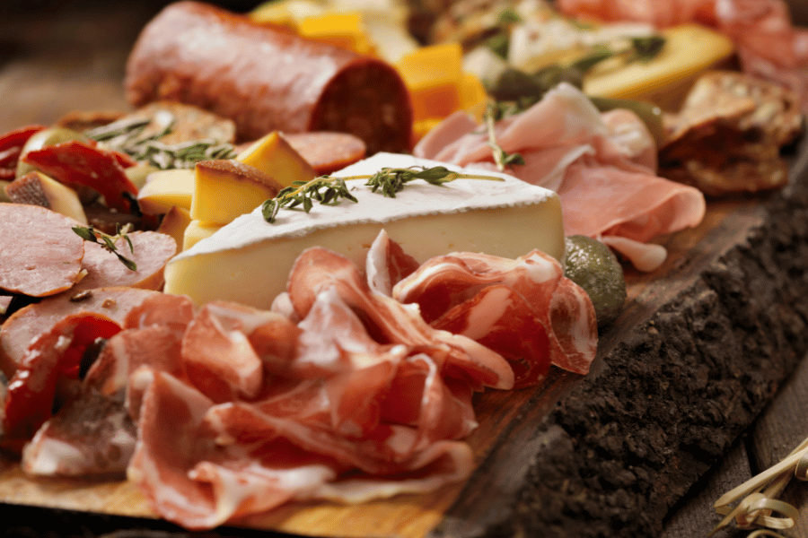 Close up image of charcuterie board with a triangle cube of cheese,proscuitto,salami,and more on a wooden board