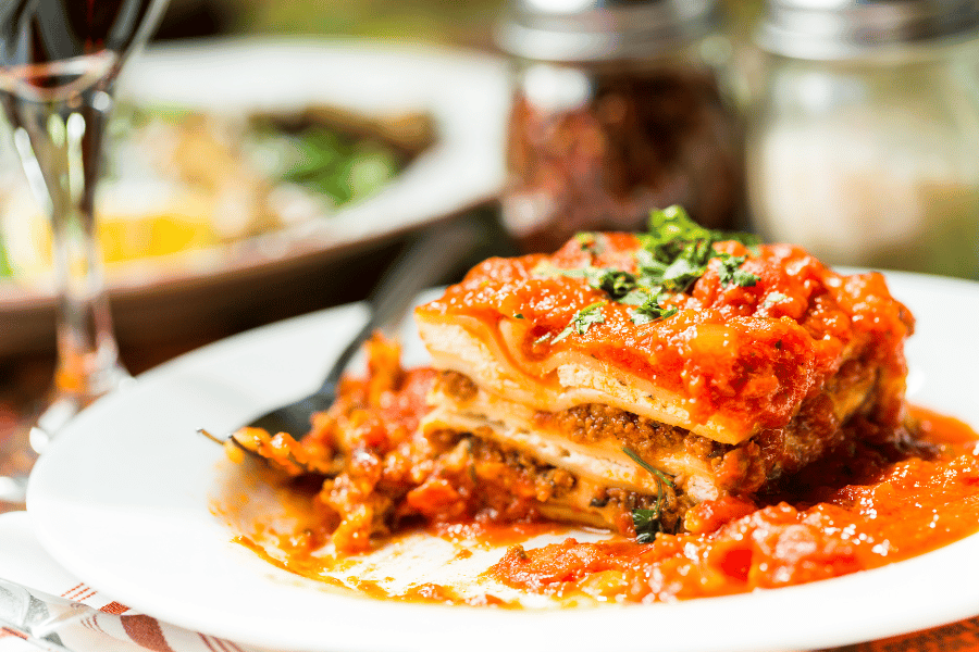 Lasagna on a white plate with tomato sauce and a wine glass in the background