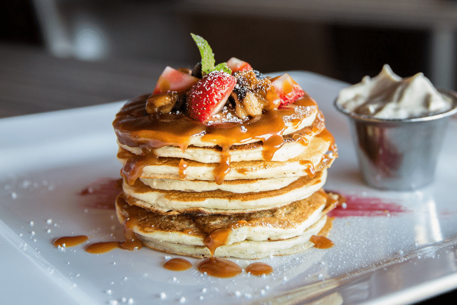 stack of pancakes on a white plate with fruit, butter, and syrup