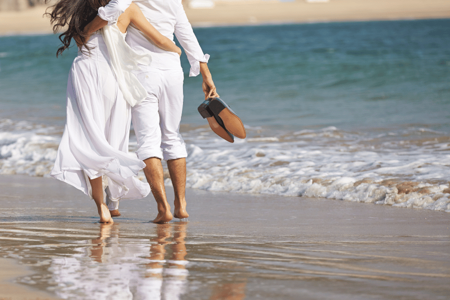 Lover Keys A photo from behind a couple dressed in all white walking in the sand along the water from shoulders down